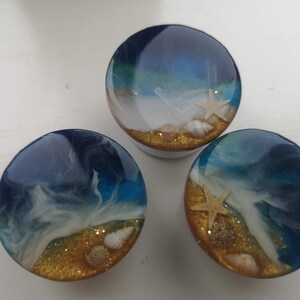 May include: Three resin phone grips with a beach design. Each round grip features a blue and white wave pattern, a golden sand base, seashells, and a starfish. The grips are on a white surface.