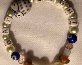 Peut inclure: Un bracelet en perles blanches avec le mot "DAYLIGHT" écrit en lettres dorées. Le bracelet comprend également un charme hibou bleu et blanc, des charmes œil bleu et des charmes champignon brun.