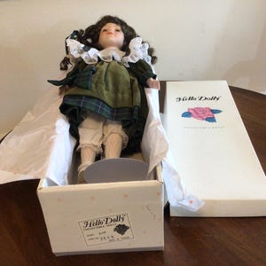 K&ouml;nnte beinhalten: Eine Sammlerpuppe in einer Box. Die Puppe hat braunes lockiges Haar, einen wei&szlig;en Spitzenkragen und ein gr&uuml;nes kariertes Kleid. Die Box ist wei&szlig; mit den Worten "Hello Dolly" und einem Rosenlogo.