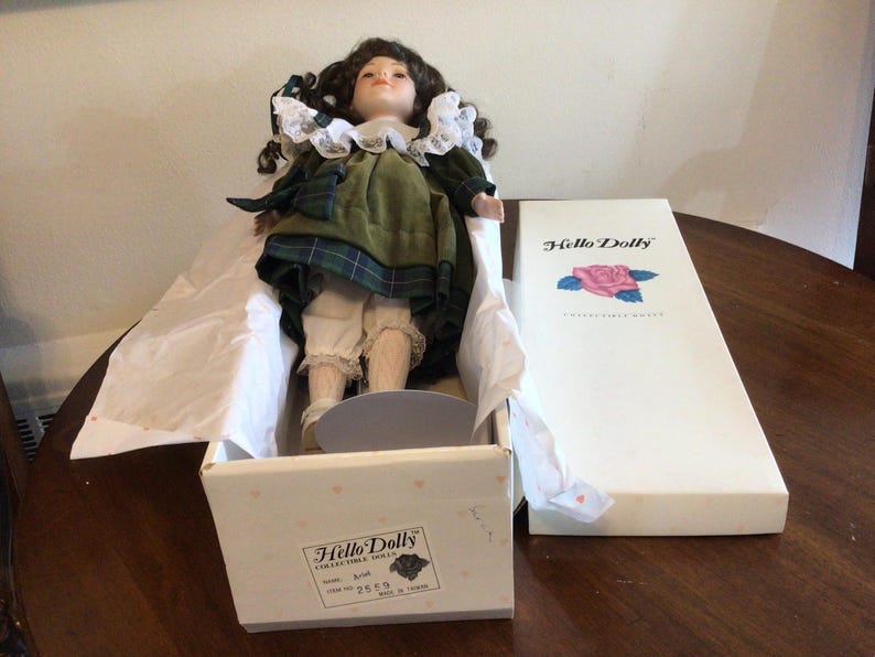 K&ouml;nnte beinhalten: Eine Sammlerpuppe in einer Box. Die Puppe hat braunes lockiges Haar, einen wei&szlig;en Spitzenkragen und ein gr&uuml;nes kariertes Kleid. Die Box ist wei&szlig; mit den Worten "Hello Dolly" und einem Rosenlogo.