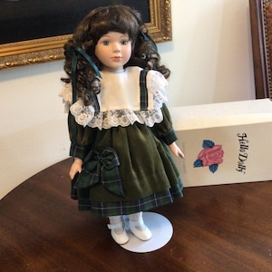 K&ouml;nnte beinhalten: Eine Vintage-Porzellanpuppe mit braunem, lockigem Haar, die ein gr&uuml;nes Samtkleid mit einer karierten Schleife und Besatz tr&auml;gt. Die Puppe hat einen wei&szlig;en Spitzkragen und steht auf einem wei&szlig;en St&auml;nder. Eine Schachtel mit der Aufschrift "Hello Dolly" befindet sich hinter der Puppe.