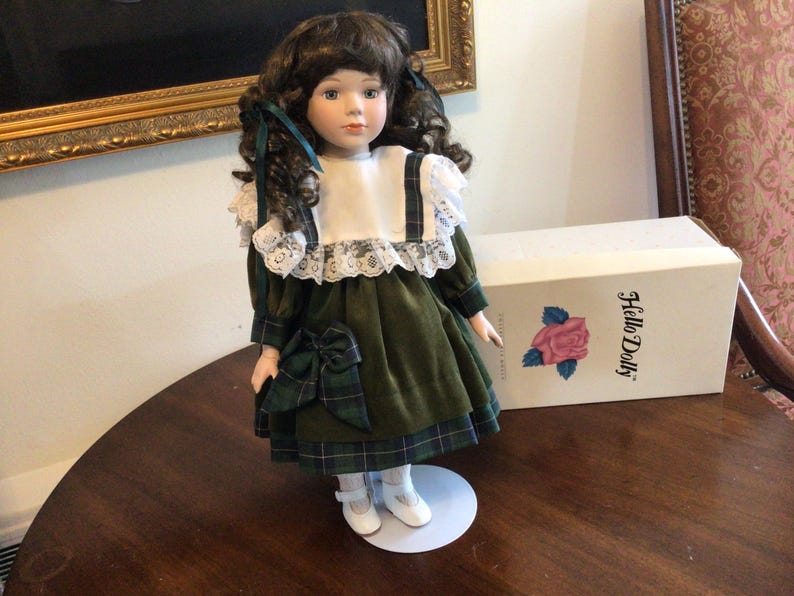 K&ouml;nnte beinhalten: Eine Vintage-Porzellanpuppe mit braunem, lockigem Haar, die ein gr&uuml;nes Samtkleid mit einer karierten Schleife und Besatz tr&auml;gt. Die Puppe hat einen wei&szlig;en Spitzkragen und steht auf einem wei&szlig;en St&auml;nder. Eine Schachtel mit der Aufschrift "Hello Dolly" befindet sich hinter der Puppe.