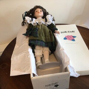 K&ouml;nnte beinhalten: Vintage Porzellanpuppe in einer Box. Die Puppe hat dunkles, lockiges Haar, ein gr&uuml;nes Kleid mit Schottenmuster-Akzenten und wei&szlig;e Spitzentrimm. Die Box ist wei&szlig; mit den Worten "Hello Dolly" und einer Rosenillustration.