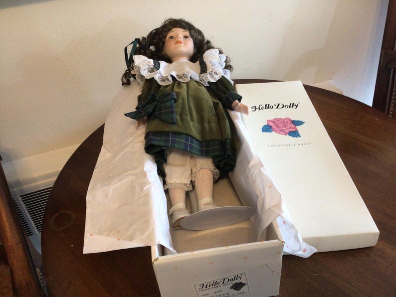 K&ouml;nnte beinhalten: Vintage Porzellanpuppe in einer Box. Die Puppe hat dunkles, lockiges Haar, ein gr&uuml;nes Kleid mit Schottenmuster-Akzenten und wei&szlig;e Spitzentrimm. Die Box ist wei&szlig; mit den Worten "Hello Dolly" und einer Rosenillustration.