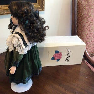 K&ouml;nnte beinhalten: Porzellanpuppe in einem gr&uuml;nen Samtkleid mit wei&szlig;em Spitzenkragen und dunklem Lockenhaar. Die Puppe steht auf einem wei&szlig;en St&auml;nder neben einer Schachtel mit der Aufschrift "Hello Dolly" und einer Rosenillustration.