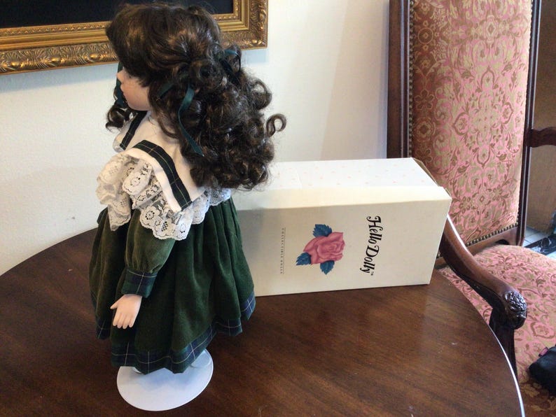 K&ouml;nnte beinhalten: Porzellanpuppe in einem gr&uuml;nen Samtkleid mit wei&szlig;em Spitzenkragen und dunklem Lockenhaar. Die Puppe steht auf einem wei&szlig;en St&auml;nder neben einer Schachtel mit der Aufschrift "Hello Dolly" und einer Rosenillustration.