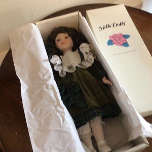 K&ouml;nnte beinhalten: Sammelpuppe "Hello Dolly" in der Originalverpackung. Die Puppe hat braunes Haar, einen wei&szlig;en Spitzenkragen und ein gr&uuml;nes Samtkleid mit Karomuster. Die Box ist wei&szlig; mit dem "Hello Dolly"-Logo und einer Rosenillustration.