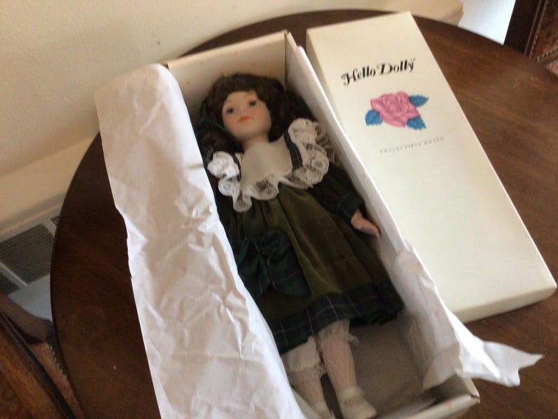 K&ouml;nnte beinhalten: Sammelpuppe "Hello Dolly" in der Originalverpackung. Die Puppe hat braunes Haar, einen wei&szlig;en Spitzenkragen und ein gr&uuml;nes Samtkleid mit Karomuster. Die Box ist wei&szlig; mit dem "Hello Dolly"-Logo und einer Rosenillustration.