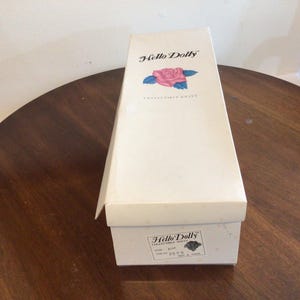 K&ouml;nnte beinhalten: Eine Vintage-Box f&uuml;r eine Hello Dolly Sammlerpuppe, mit den Worten "Hello Dolly" und einer Rosengrafik auf dem Deckel. Die Box steht auf einem dunklen Holztisch.