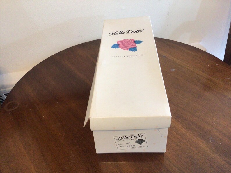 K&ouml;nnte beinhalten: Eine Vintage-Box f&uuml;r eine Hello Dolly Sammlerpuppe, mit den Worten "Hello Dolly" und einer Rosengrafik auf dem Deckel. Die Box steht auf einem dunklen Holztisch.