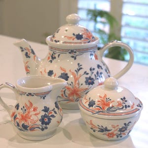 May include: A white ceramic tea set featuring a teapot, creamer jug, and sugar bowl. Each piece is adorned with a floral design in blue and orange. The teapot has a lid and a handle. The creamer has a handle and a spout. The sugar bowl has a lid.