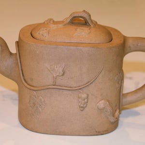 May include: A light brown, cylindrical clay teapot featuring a textured surface with relief carvings of grapevines, leaves, and clusters of grapes. The teapot includes a matching lid with a small handle, a curved spout, and a sturdy side handle.