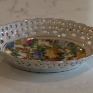 K&ouml;nnte beinhalten: Eine wei&szlig;e Keramikschale mit einem Blumendesign und einem spitzengleichen Muster am Rand. Die Schale ist oval geformt und hat an jeder Seite einen kleinen Griff.