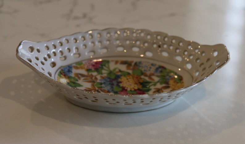 K&ouml;nnte beinhalten: Eine wei&szlig;e Keramikschale mit einem Blumendesign und einem spitzengleichen Muster am Rand. Die Schale ist oval geformt und hat an jeder Seite einen kleinen Griff.