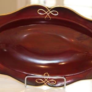 May include: A burgundy oval serving dish with a gold rim and intricate gold designs. The dish is made of ceramic and has a glossy finish.
