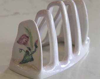 May include: A white ceramic toast rack with four slots. The side features a hand-painted floral design with purple flowers, green leaves, and stems. The rack is designed to hold multiple slices of toast and has a curved handle.
