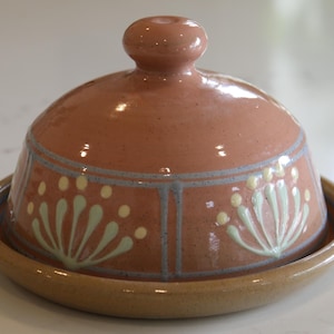 May include: A terracotta ceramic butter dish with a domed lid and a round base. The dish features a floral design in light green and yellow. The lid has a small knob. The dish is suitable for storing butter.