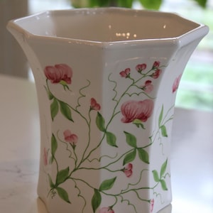 May include: White ceramic vase with a hexagonal design, featuring hand-painted pink flowers and green foliage. The vase has a flared top and a smooth, glossy finish, ideal for displaying flowers or as a decorative item.