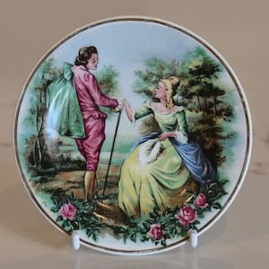 May include: A vintage ceramic plate with a painted scene of a couple in a garden setting. The man is wearing a pink jacket and the woman is wearing a yellow and blue dress. The plate has a gold rim and is decorated with roses.