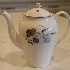 May include: White ceramic coffee pot with a floral design featuring brown, gray, and white leaves and a white flower. The pot has a gold lid and a gold trim around the rim.