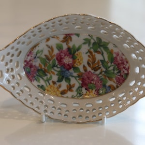K&ouml;nnte beinhalten: Eine wei&szlig;e ovale Porzellansch&uuml;ssel mit Blumenmuster. Die Sch&uuml;ssel hat einen spitzengleichen Rand und einen goldenen Rand.