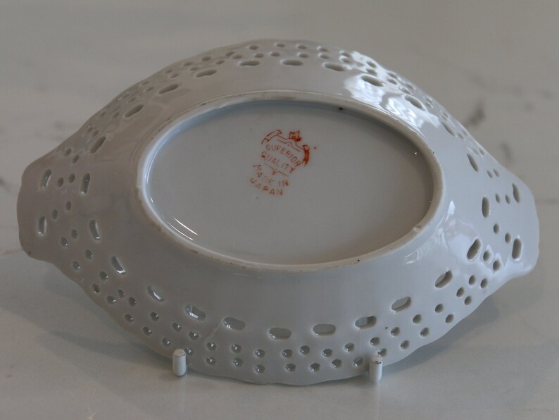 K&ouml;nnte beinhalten: Wei&szlig;e Keramik-Servierplatte mit gewelltem Rand und einem spitzengleichen Design. Die Platte hat eine kleine ovale Form in der Mitte mit dem Text "Superior Quality Made in Japan" in Rot.
