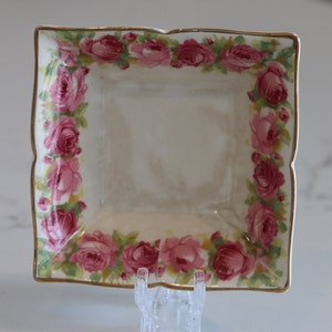May include: A small, square, white porcelain dish with a gold rim and a pink rose floral pattern.