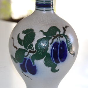May include: A ceramic vase with a speckled, light grey body. The vase features a design of green leaves and dark blue plums. The neck of the vase is dark blue and green.