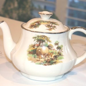 May include: A cream-coloured ceramic teapot featuring a silver-trimmed rim, spout, and handle. The body and lid are decorated with a colourful pastoral scene depicting a horse-drawn hay cart in a field with trees, a fence, and a small cottage.