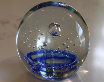 May include: A clear glass paperweight with a blue spiral design and small air bubbles suspended within. The paperweight is spherical and reflects light, creating a shimmering effect. The blue spiral is at the bottom of the paperweight.