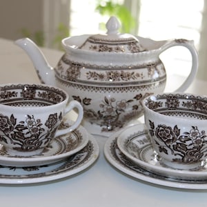 May include: A white tea set with a brown floral design. The set comprises a teapot, two teacups, and saucers. The teapot has a lid and a handle. The teacups and saucers are circular. The set is displayed on a white surface.