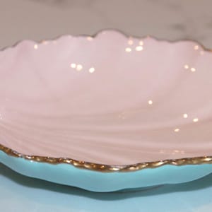 May include: A shell-shaped ceramic dish featuring a smooth pale pink interior and a vibrant turquoise exterior. The scalloped edges of the dish are finished with a delicate gold-toned trim, highlighting the curved, organic form of the piece.