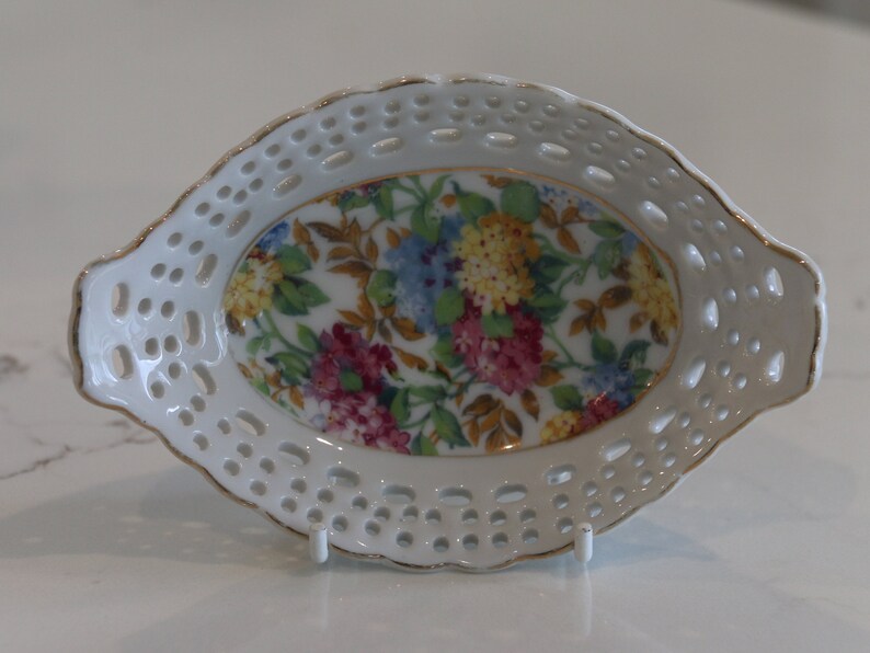 K&ouml;nnte beinhalten: Wei&szlig;e ovale Porzellansch&uuml;ssel mit Blumenmuster und einer spitzengleichen Bord&uuml;re. Die Sch&uuml;ssel hat einen goldenen Rand.