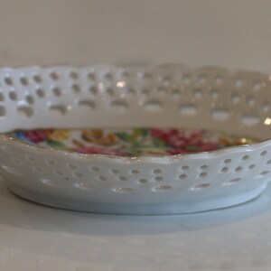 K&ouml;nnte beinhalten: Eine wei&szlig;e Porzellansch&uuml;ssel mit einem spitzengleichen Design und einem Blumenmuster auf dem Boden. Die Sch&uuml;ssel hat zwei Henkel.