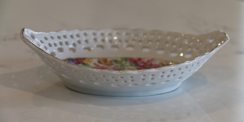 K&ouml;nnte beinhalten: Eine wei&szlig;e Porzellansch&uuml;ssel mit einem spitzengleichen Design und einem Blumenmuster auf dem Boden. Die Sch&uuml;ssel hat zwei Henkel.