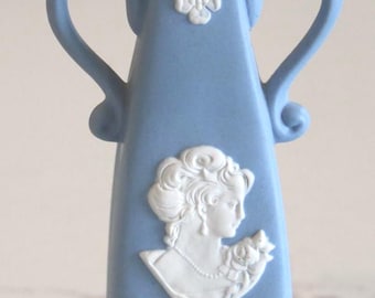 May include: A light blue vase featuring white relief decorations. The design includes a central cameo of a person in profile, a floral garland at the base, a small flower motif on the neck, and two curved handles on the sides of the vessel.