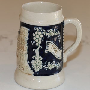 May include: A white ceramic beer stein with a blue and gold design. The stein features a depiction of a building, grapes, and a banner that reads "Trier".