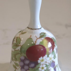 May include: A white porcelain bell featuring a colourful botanical illustration of various fruits including red plums, green pears, and purple grapes, accented with small white flowers and green leaves, finished with a thin metallic gold rim at the base.