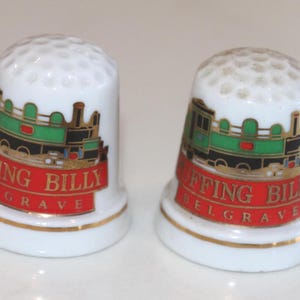 May include: Two white ceramic thimbles featuring a dimpled top and a gold-trimmed base. Each thimble displays a colourful illustration of a green and black steam locomotive above a red banner with the gold text PUFFING BILLY and BELGRAVE.