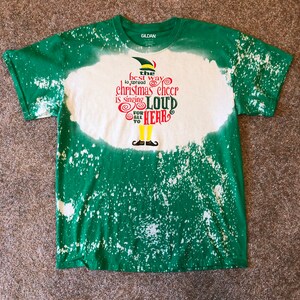 May include: Green and white tie-dye t-shirt with a bleach-splattered design. The shirt features a cartoon elf with the text "the best way to spread christmas cheer is singing loud for all to hear".