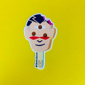 May include: A sticker of a cartoon face with a third eye, red and yellow accents, and a pink streak in the hair. The face is on a popsicle stick with the text "@Buena Biscuits" and a bunny rabbit logo.