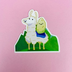 May include: A white llama with a brown potato on its back. The llama is standing on a green grassy hill with mountains in the background.