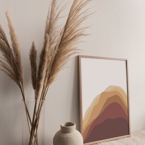Puede incluir: Una impresi&oacute;n art&iacute;stica enmarcada con un dise&ntilde;o de monta&ntilde;a abstracto en tonos marrones y amarillos. Un jarr&oacute;n de vidrio transparente contiene hierba de la pampa seca, y un peque&ntilde;o jarr&oacute;n de cer&aacute;mica beige est&aacute; cerca. Los objetos est&aacute;n dispuestos sobre una superficie tejida.