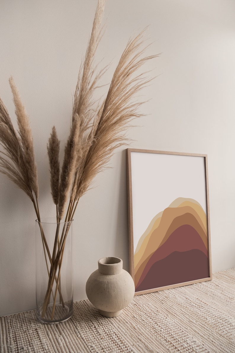 Puede incluir: Una impresi&oacute;n art&iacute;stica enmarcada con un dise&ntilde;o de monta&ntilde;a abstracto en tonos marrones y amarillos. Un jarr&oacute;n de vidrio transparente contiene hierba de la pampa seca, y un peque&ntilde;o jarr&oacute;n de cer&aacute;mica beige est&aacute; cerca. Los objetos est&aacute;n dispuestos sobre una superficie tejida.