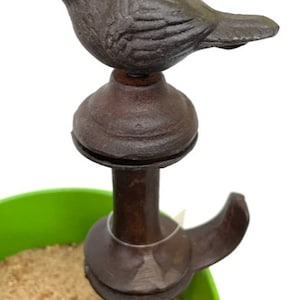 May include: A dark brown cast iron bird garden stake. The bird is perched atop a decorative stake with a hook. The stake is placed in a green container filled with sand.