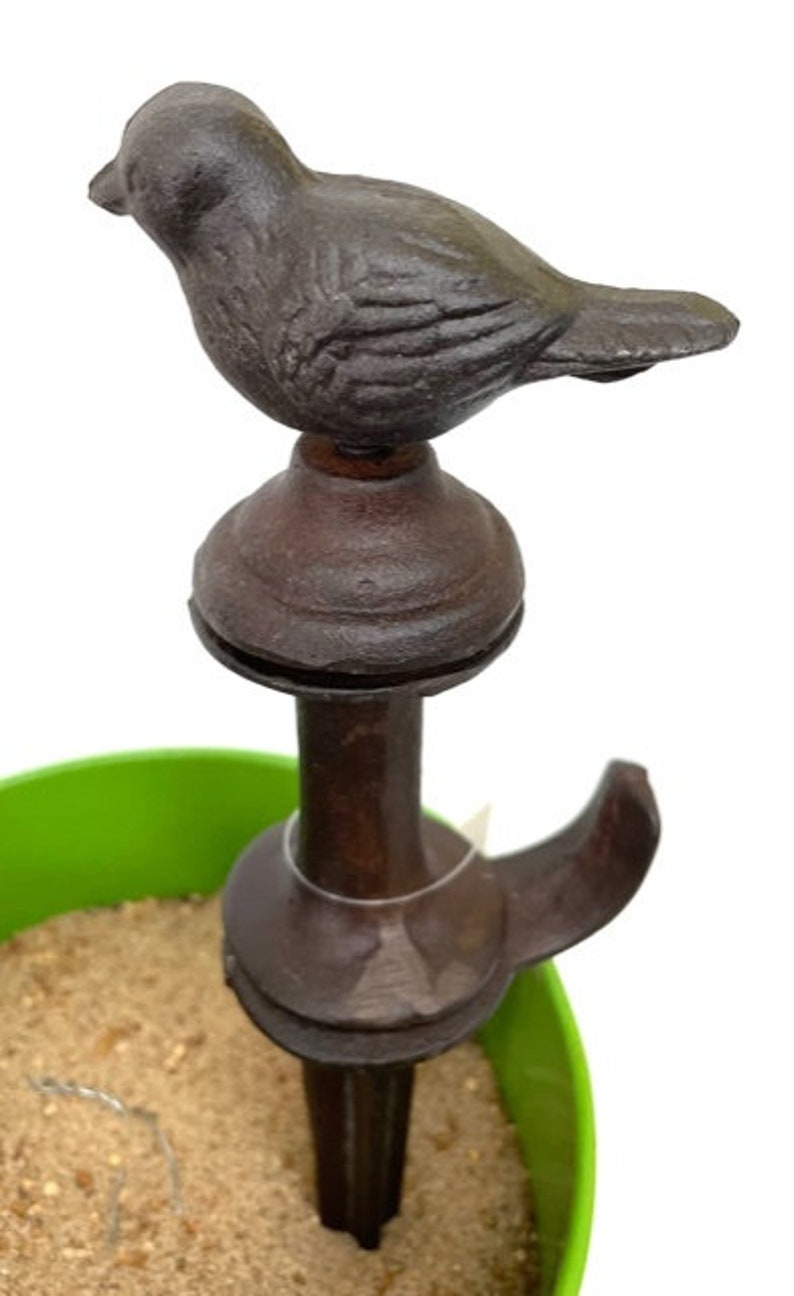 May include: A dark brown cast iron bird garden stake. The bird is perched atop a decorative stake with a hook. The stake is placed in a green container filled with sand.