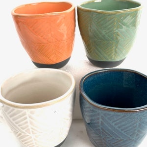 May include: Four ceramic mugs with different colored glazes and geometric patterns. The mugs are all the same size and shape, and they have a black rim.