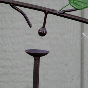Pu&ograve; includere: Un'alimentatore decorativo in metallo marrone scuro con un ramo, una foglia e un piccolo elemento rotondo sospeso. L'alimentatore ha una piccola base rotonda. Una mano tiene una corda bianca.
