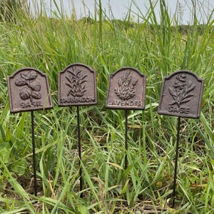 May include: Four weathered metal garden markers with plant illustrations and names: Basil, Tarragon, Lavender, and Sage. The markers are brown with black stems, set in a grassy outdoor environment.