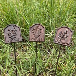 May include: Three weathered metal garden markers with the names "Thyme", "Chives", and "Rosemary" embossed on them. Each marker features a different embossed herb illustration. The markers are set in a grassy area.